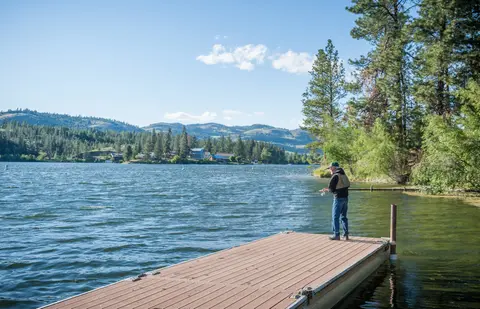 A person fishes from a wooden dock on a lake with distant hills and trees.
