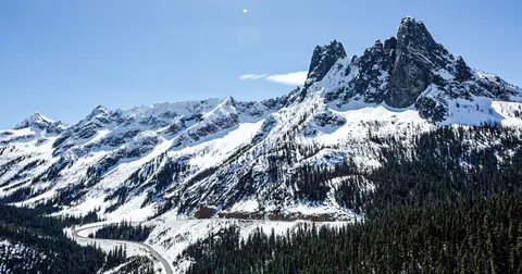 A scenic landscape featuring snow-covered mountains, a winding road, and a dense pine forest.