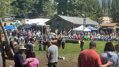 An outdoor gathering of people watching an event with logs, tents, and buildings in the background.