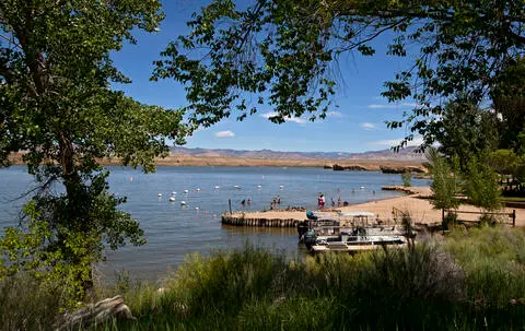 A scenic view of a lake with a sandy beach, dock, and distant hills framed by trees.