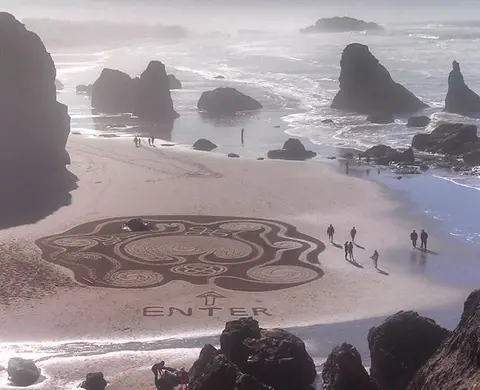 A large sand labyrinth drawn on a beach with ocean waves and rock formations in the background.