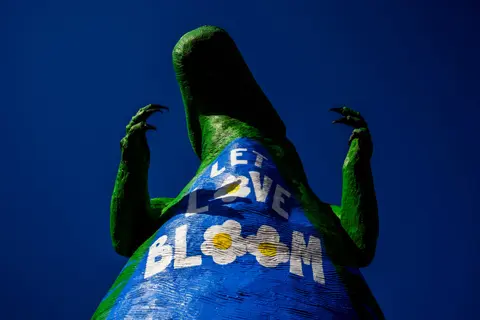 A low-angle view of a large dinosaur statue painted with a message and flowers against a blue sky.