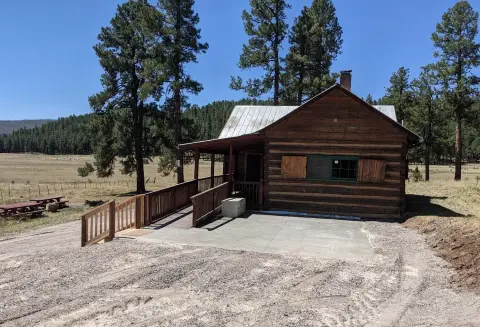A rustic wooden cabin with a porch and ramp sits in a grassy field surrounded by trees.