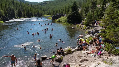 People swimming and relaxing in a scenic river surrounded by a forest and mountains.