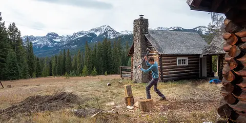 A person chops wood outside a rustic cabin with snowy mountains in the background.