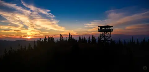 A fire lookout tower stands silhouetted against a colorful sunset sky over a forest ridge.