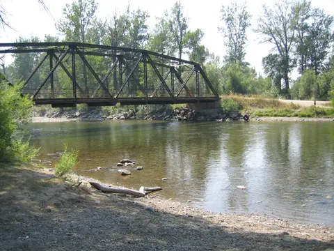 A metal truss bridge spans over a calm river surrounded by trees.