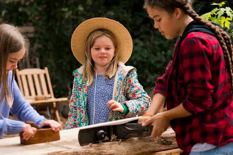 Three young people work with woodworking tools outdoors.