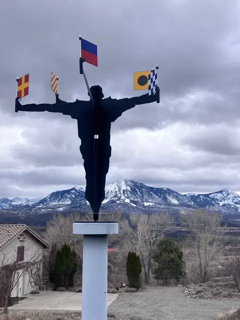 A silhouette sculpture holding flags stands outdoors with mountains in the background.