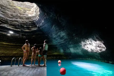 A family stands on a wooden dock inside a cave with a pool of water and an opening to the sky above.