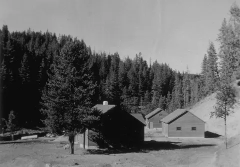Black and white photograph showing a group of wooden buildings nestled in a forested mountainous area.