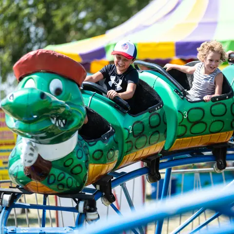 Two children riding a green alligator-themed roller coaster at a fair.