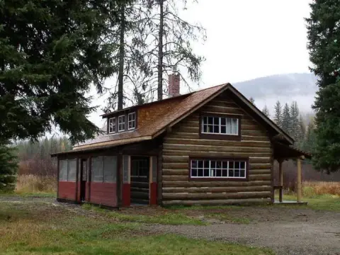 A log cabin sits in a clearing surrounded by trees and mountains.