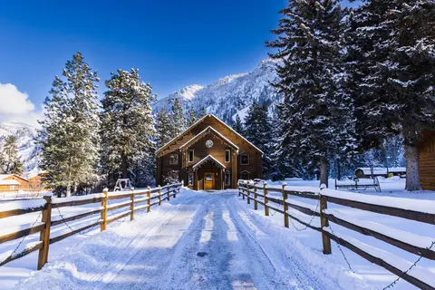 A wooden building stands at the end of a snow-covered path flanked by fences and pine trees against a mountain backdrop.