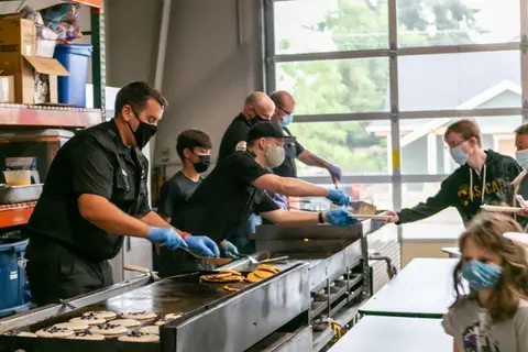 People wearing masks cook and serve pancakes from large griddles indoors.