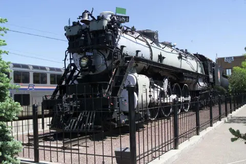A large vintage steam locomotive is displayed behind a black metal fence outdoors.