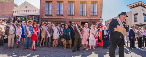 A man speaks into a microphone before a group of people dressed in historical costumes on a street.