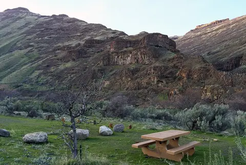 A picnic table sits in a grassy area with sparse vegetation before large rocky hills and mountains.