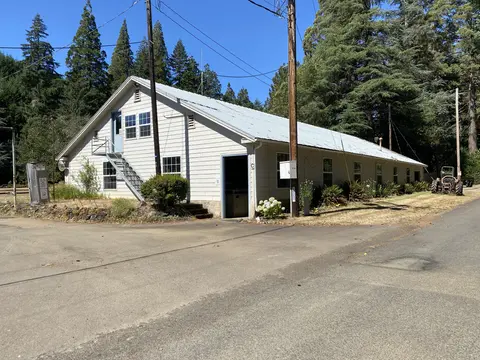 A long, light-colored building with a metal roof stands among tall trees.