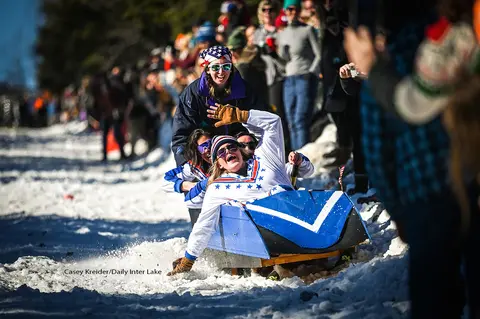 People riding a homemade sled down a snow-covered hill in front of a crowd.