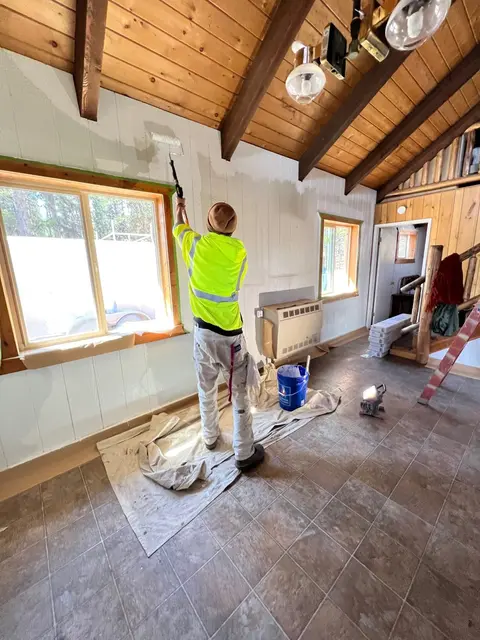 A person paints an interior wall in a room with a wooden ceiling.