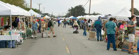 People browse and walk along a street lined with vendor tents at an outdoor market.