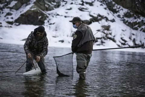 Two people in waders fish in a snowy river.