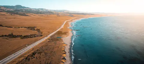 An aerial view of a coastal highway running alongside the ocean and rolling hills.