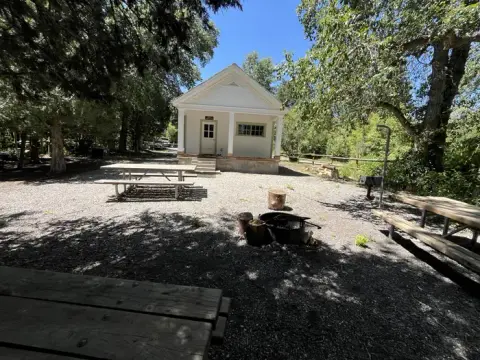 A small white building stands outdoors surrounded by trees and picnic tables.