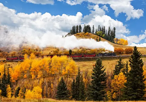 A steam train travels through a mountainous landscape with autumn foliage.