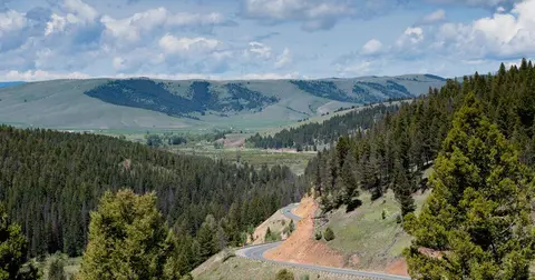 A winding road descends through a forested mountain valley toward distant hills under a cloudy sky.