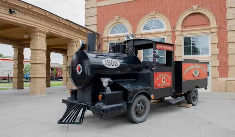 A truck modified to look like a steam locomotive parked in front of a brick building.