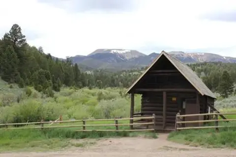 A wooden cabin stands in a grassy valley with mountains in the background.