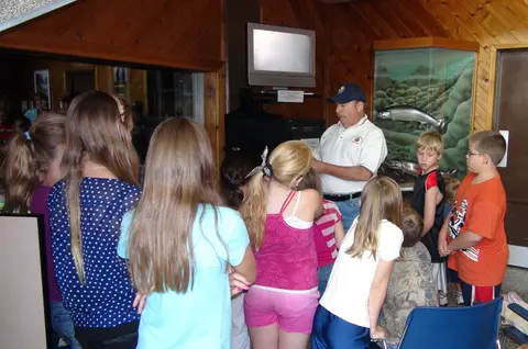A group of children listening to a speaker inside a room with wood paneling.
