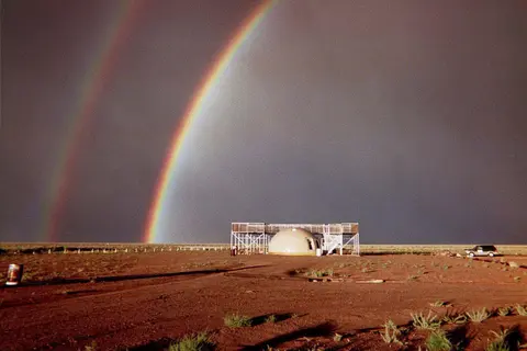 A double rainbow appears in the sky above a small dome structure on barren ground.