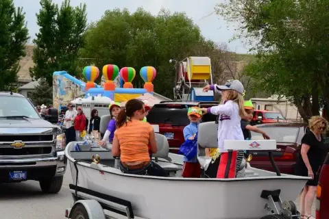 People ride in a boat on a trailer during an outdoor event with a bouncy house and carnival rides in the background.