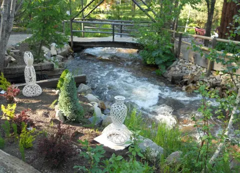 A small footbridge crosses a rushing stream in a garden with decorative white pottery sculptures.