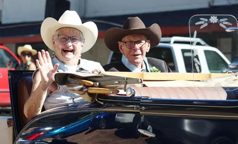 An older couple wearing cowboy hats ride in a vintage car.