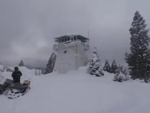 A fire lookout tower surrounded by snow with a person and snowmobiles in the foreground.