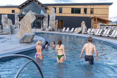 People enjoy swimming in a modern outdoor pool with large decorative rocks and a building in the background.