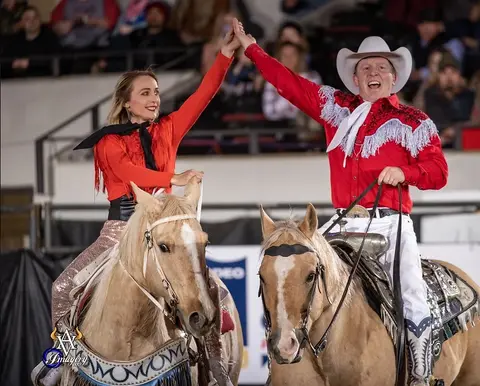 Two performers in red western attire ride horses and hold hands in an arena.