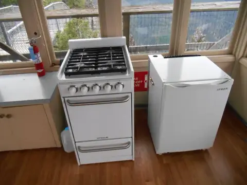 A white gas stove and a small white refrigerator sit inside a building with large windows overlooking a landscape.