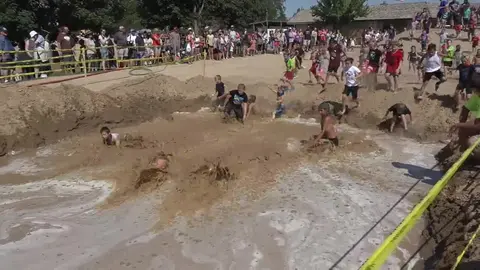 Children participate in a mud obstacle course as a crowd watches from behind a barrier.