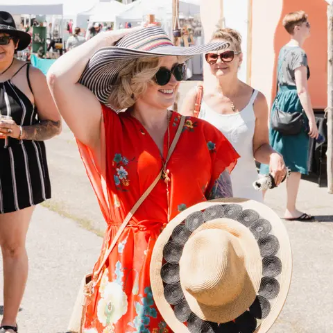 A woman wearing a large striped sunhat and red floral dress stands outdoors among other people.