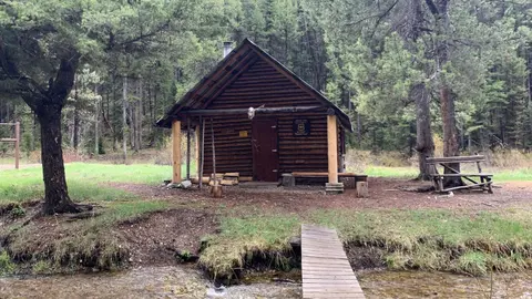 A wooden cabin in a forest with a small wooden bridge crossing a stream in front of it.
