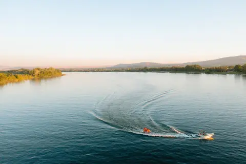 A boat tows a raft on a wide river surrounded by trees and hills.