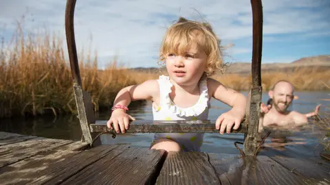 A young child climbs out of a natural pool using a metal ladder attached to a wooden dock.