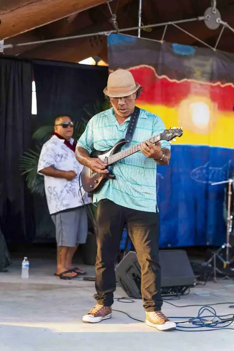 A musician plays guitar on a stage with a person in the background.