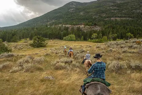 People horseback riding across a grassy meadow toward a mountain.