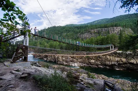 A suspension bridge spans a river with forested mountains in the background.
